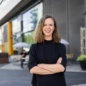 Womanwith briwn shoulder length hair in dark sweater with arms crossed standing in front of blurren background of a building.